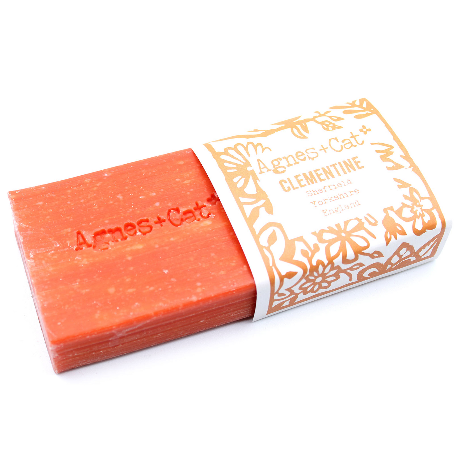 Jabón Artesanal Agnes+Cat Clementina 140g, frescura cítrica y energizante, mostrando la pastilla de jabón saliendo de su envoltorio, perfecta para una limpieza revitalizante con fragancia fresca